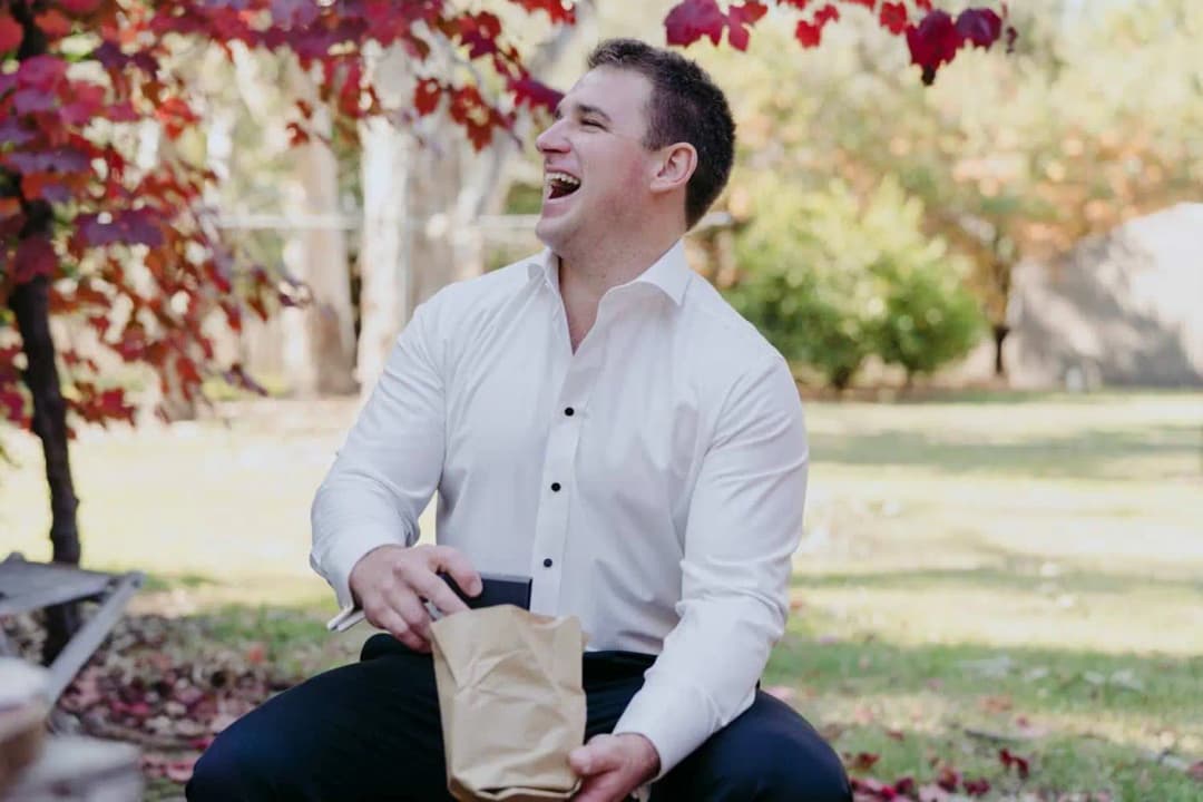 Groom laughing while holding a paper bag under a leafy pergola.