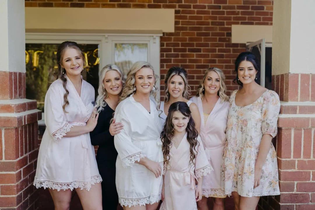 Bride in a white robe posing with five women and a young girl by a brick doorway.