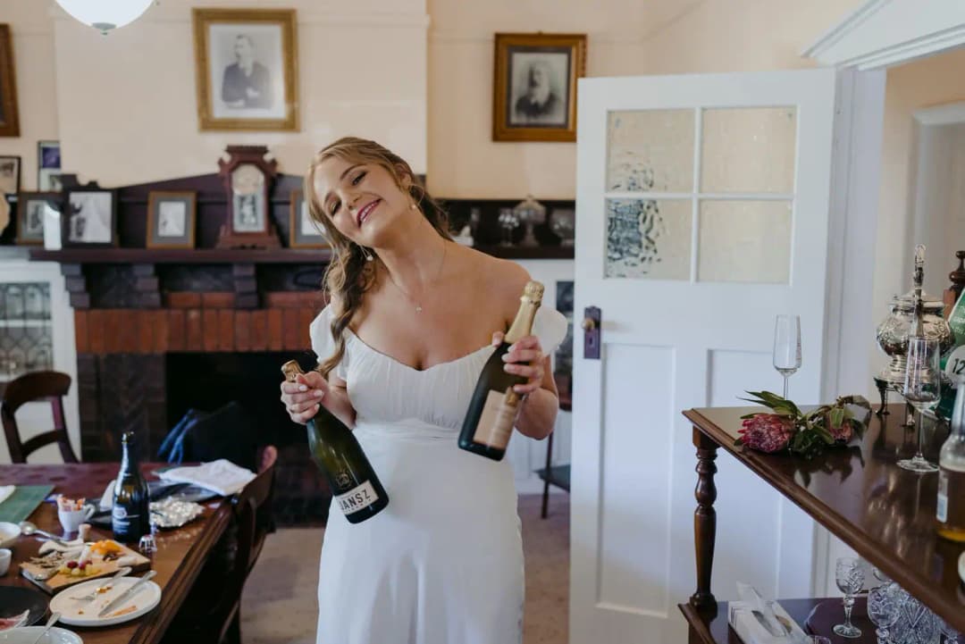 Woman in a white dress smiling while holding two sparkling wine bottles.