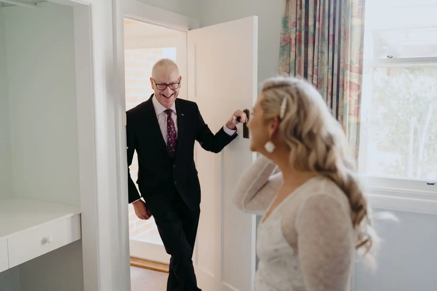 Man in glasses entering the room and smiling at the bride in her wedding dress.