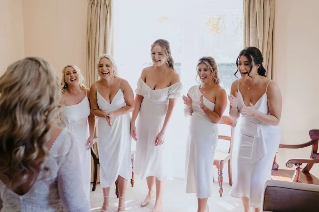 Five women in white dresses smiling at the bride inside a bright room.