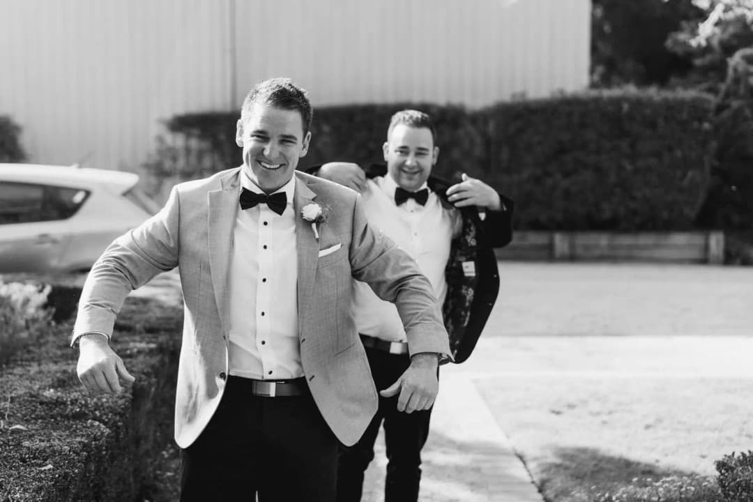 Black-and-white photograph of the groom walking outdoors while another man follows.