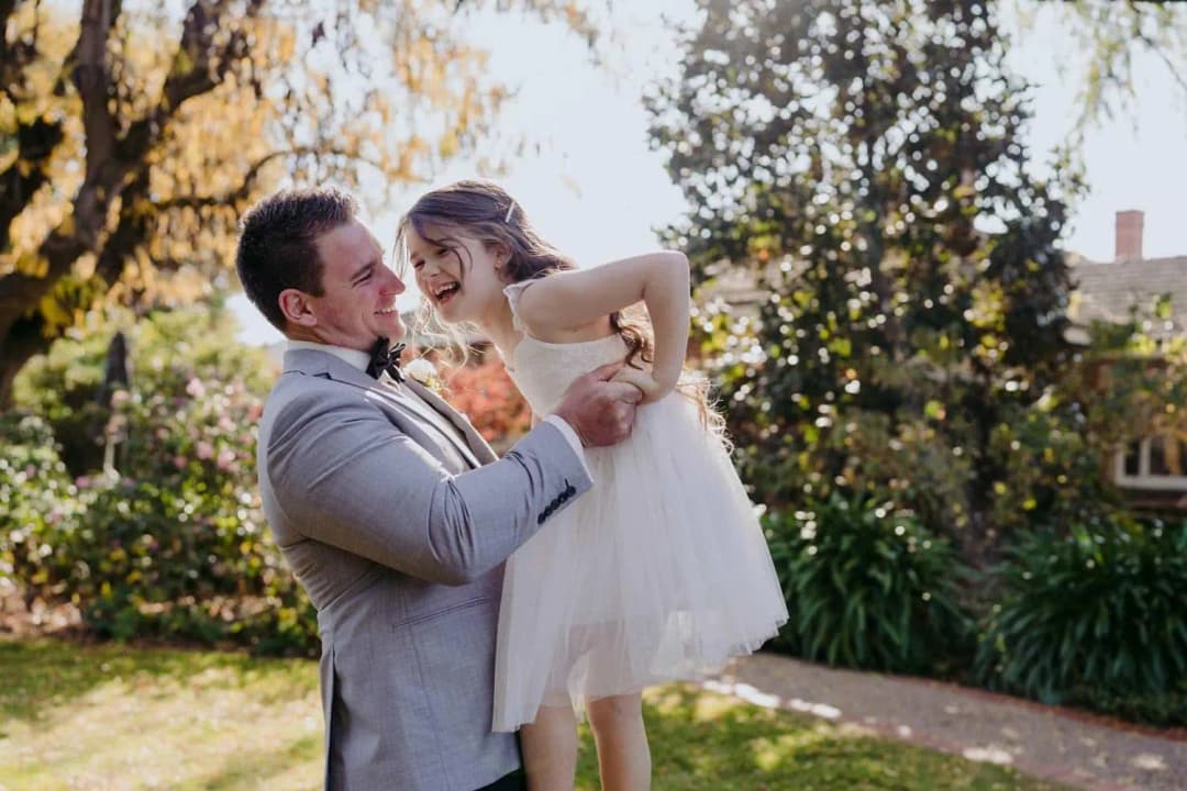 Groom lifting a smiling young girl in a white dress on a garden lawn.