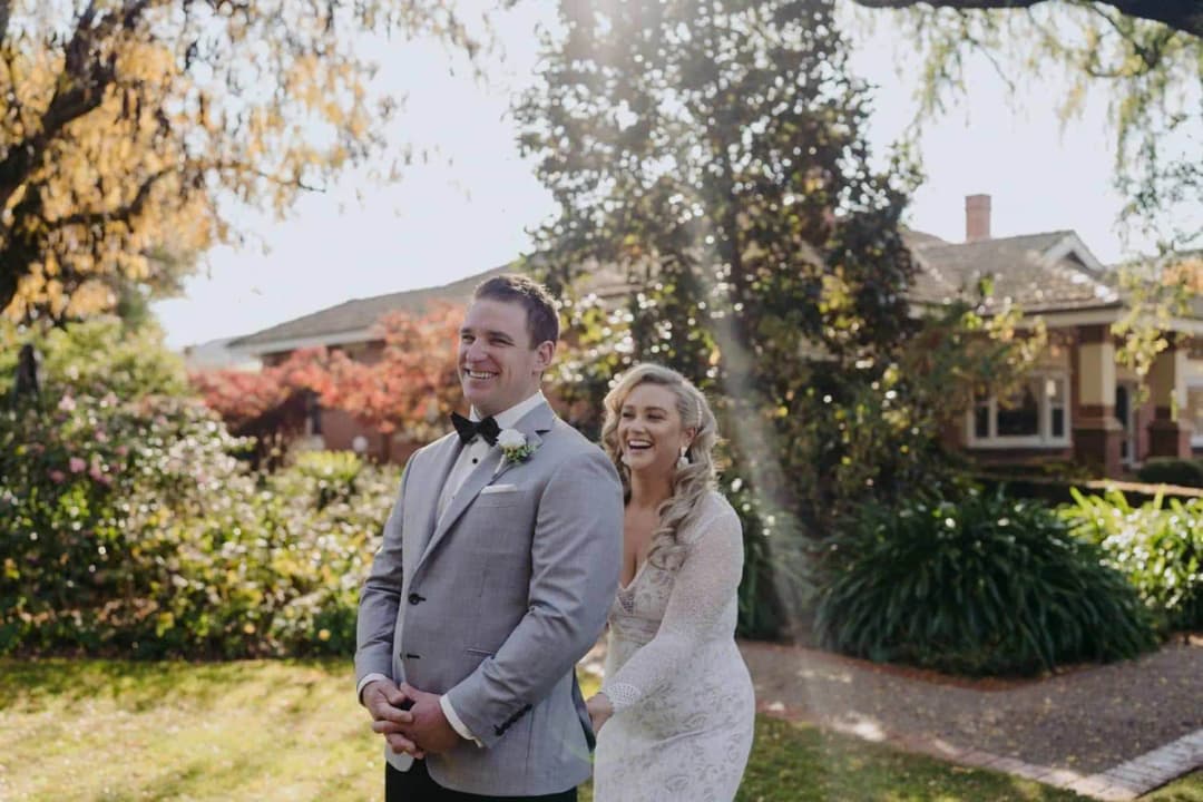 Bride approaching the groom from behind beneath sunlit garden trees.