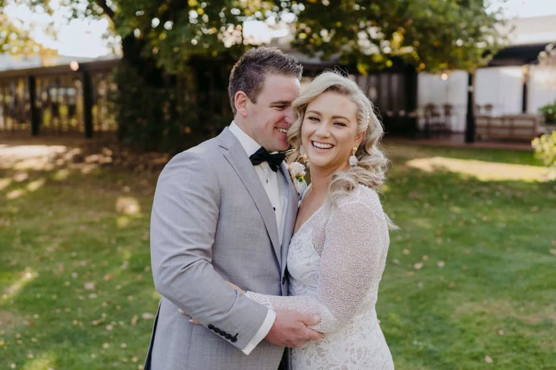 Groom leaning close to the smiling bride beneath garden trees.