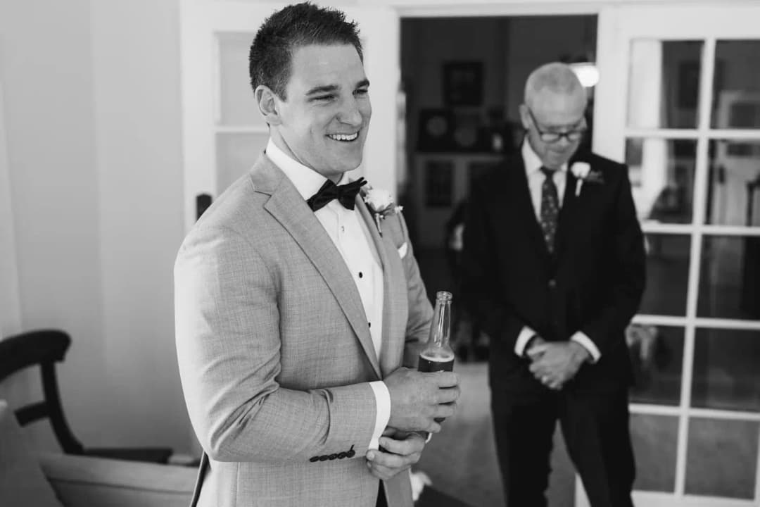 Black-and-white photograph of the smiling groom holding a drink inside a house.