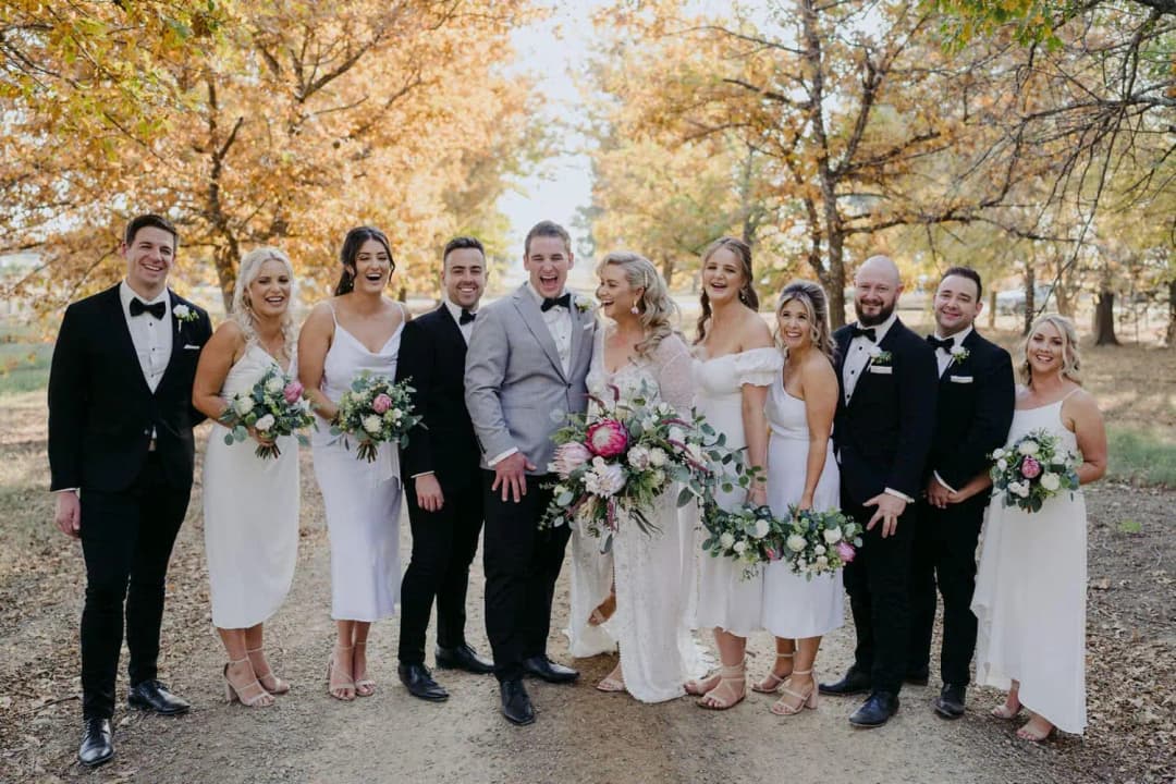 Bride and groom posing with their wedding party beneath autumn foliage.