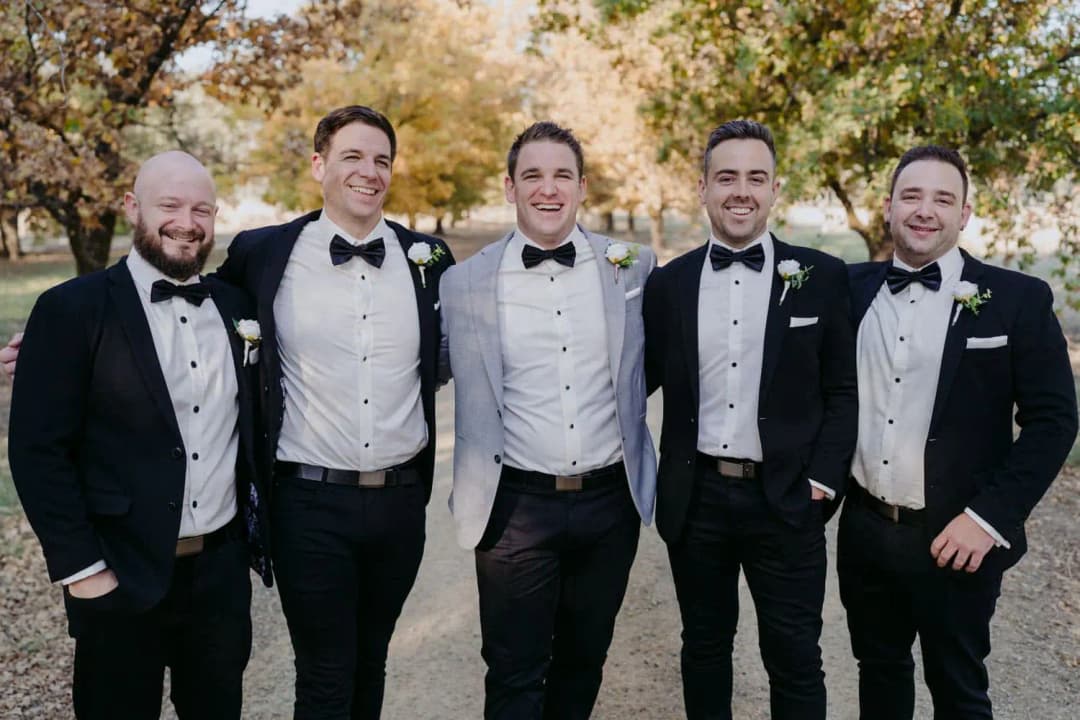 Groom and four men posing together in bow ties beneath autumn trees.