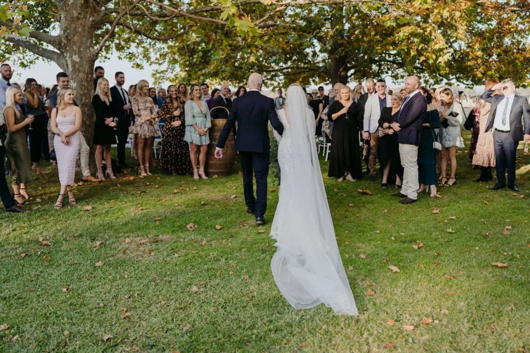 Bride and a man in a dark suit walking down the aisle beneath autumn trees.