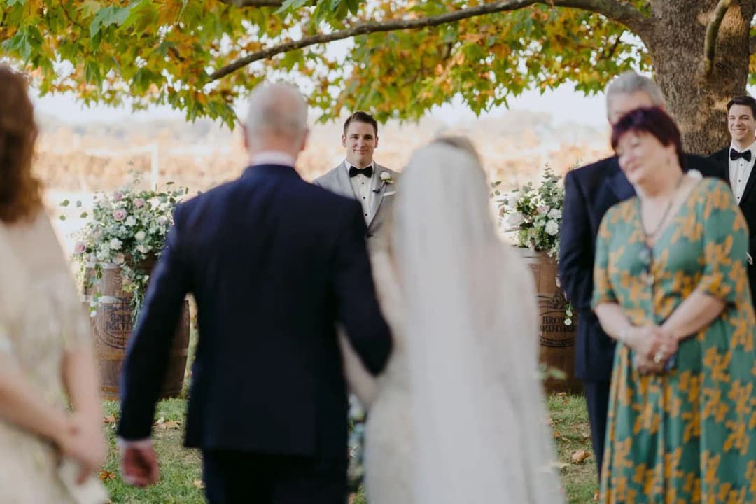 Groom watching the bride approach with a man in a dark suit.