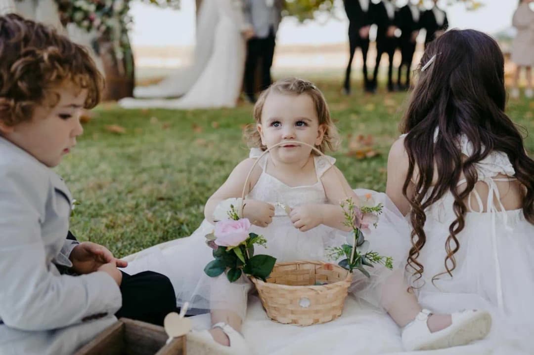 Three children sitting on a white blanket with a small flower basket.