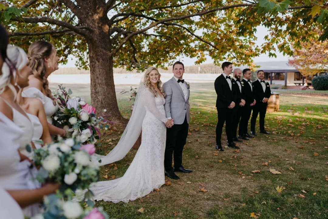 Bride and groom smiling beside their wedding party under autumn branches.