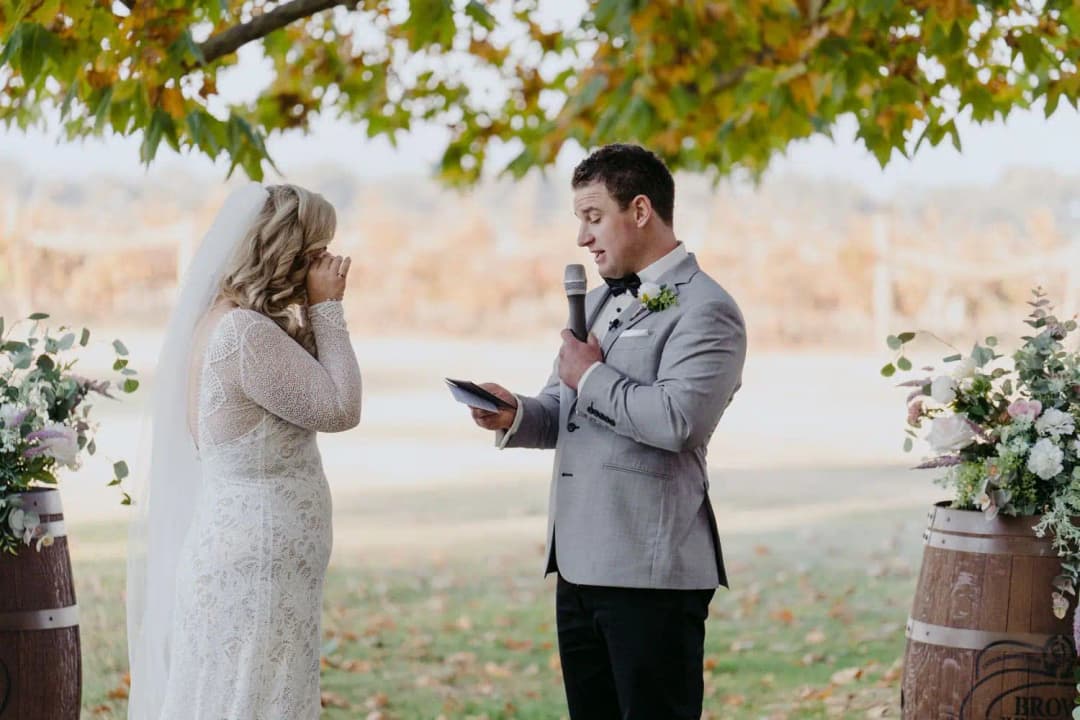 Groom reading from a phone into a microphone as the bride touches her face.