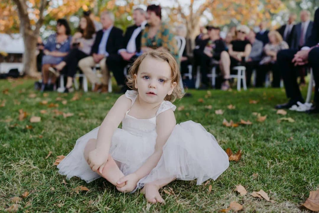 Young girl in a white tulle dress sitting on the grass in front of ceremony guests.