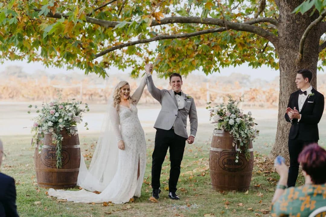 Bride and groom raising their joined hands between flower-topped wooden barrels.