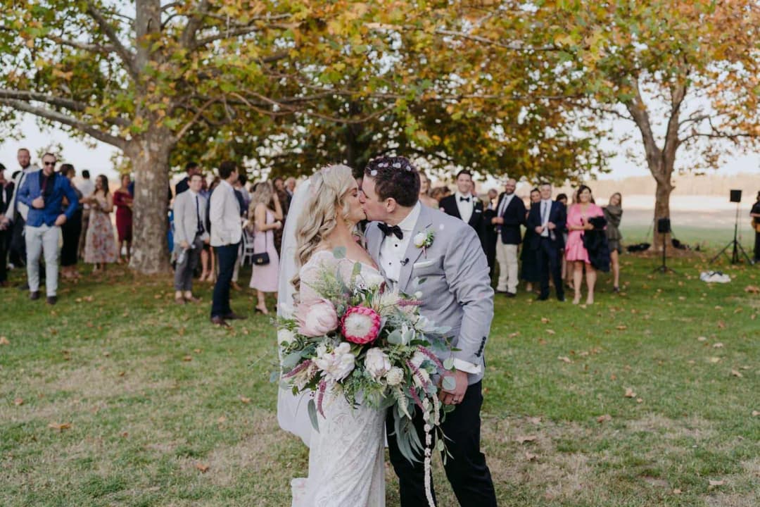 Bride and groom kissing with their bouquet while guests stand behind them.
