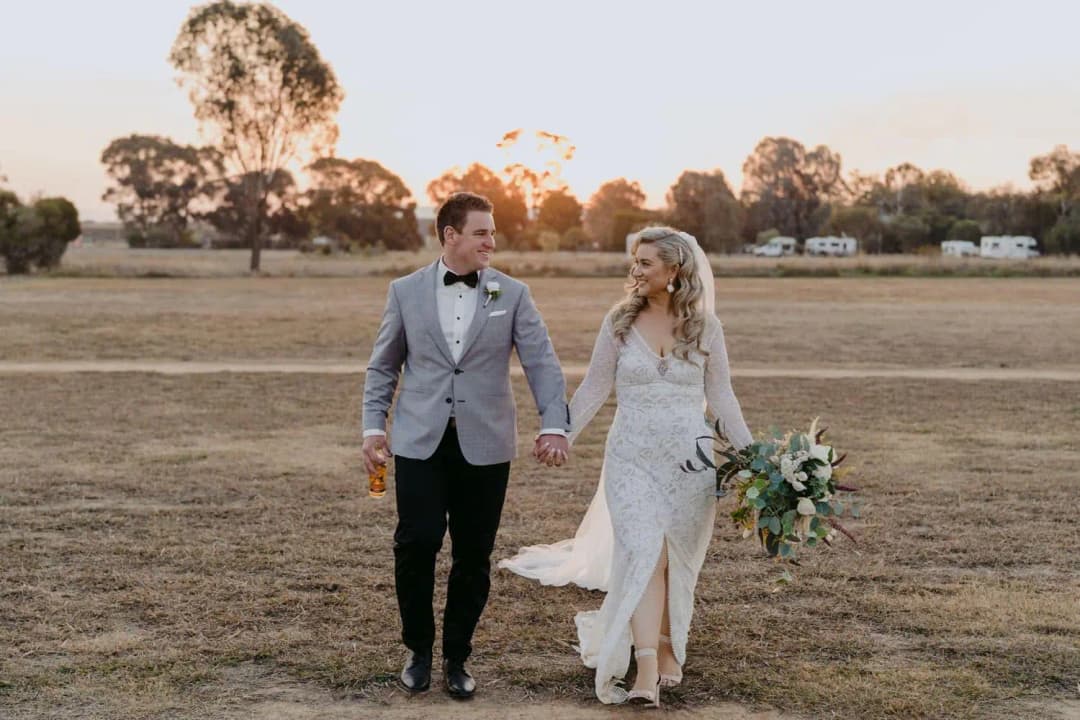 Bride and groom walking hand in hand across a dry field at sunset.