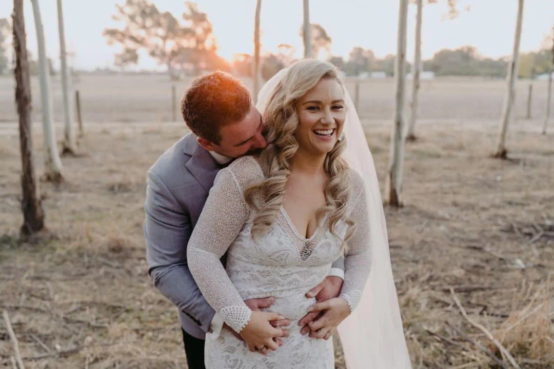 Groom embracing the laughing bride from behind among tall trees.