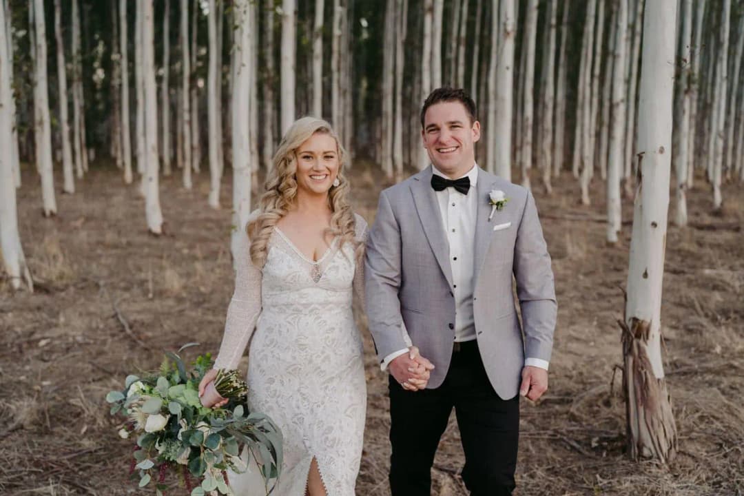 Bride and groom smiling and holding hands among rows of slender trees.