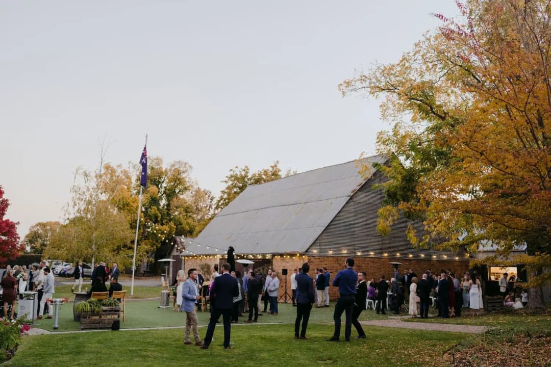 Guests gathering outside a rustic reception building beside autumn trees.