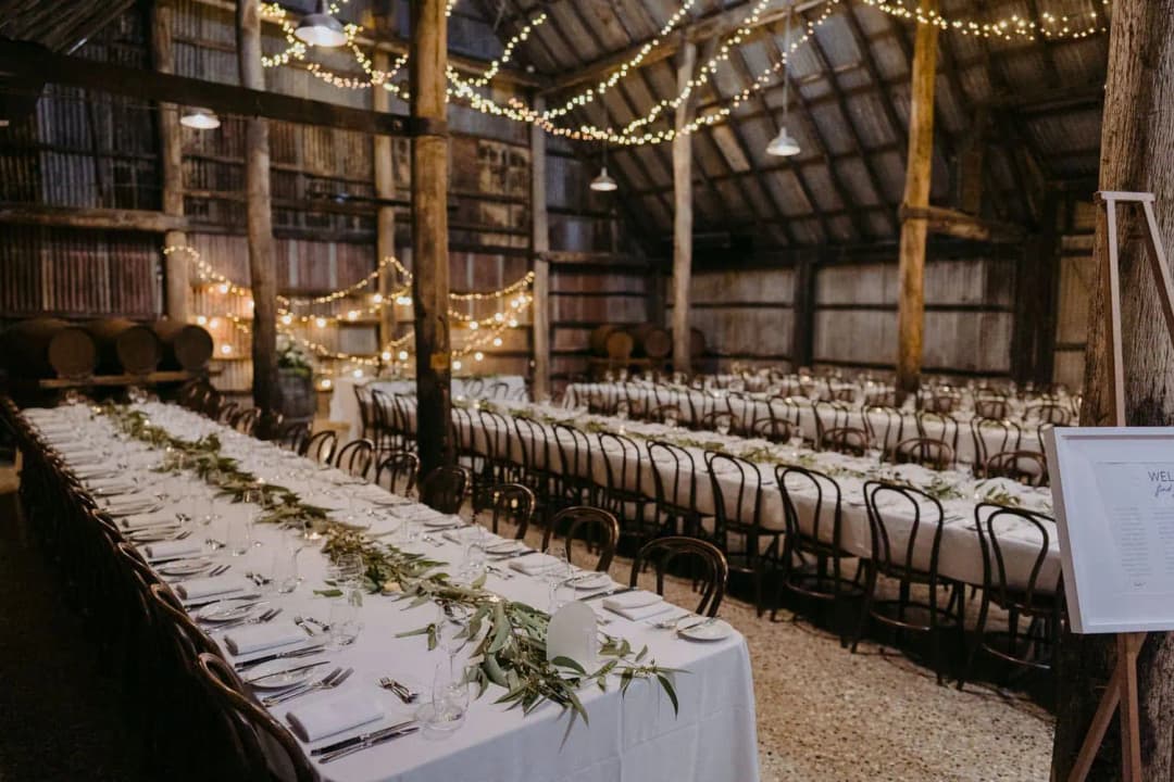 Long white reception tables beneath string lights and timber beams.