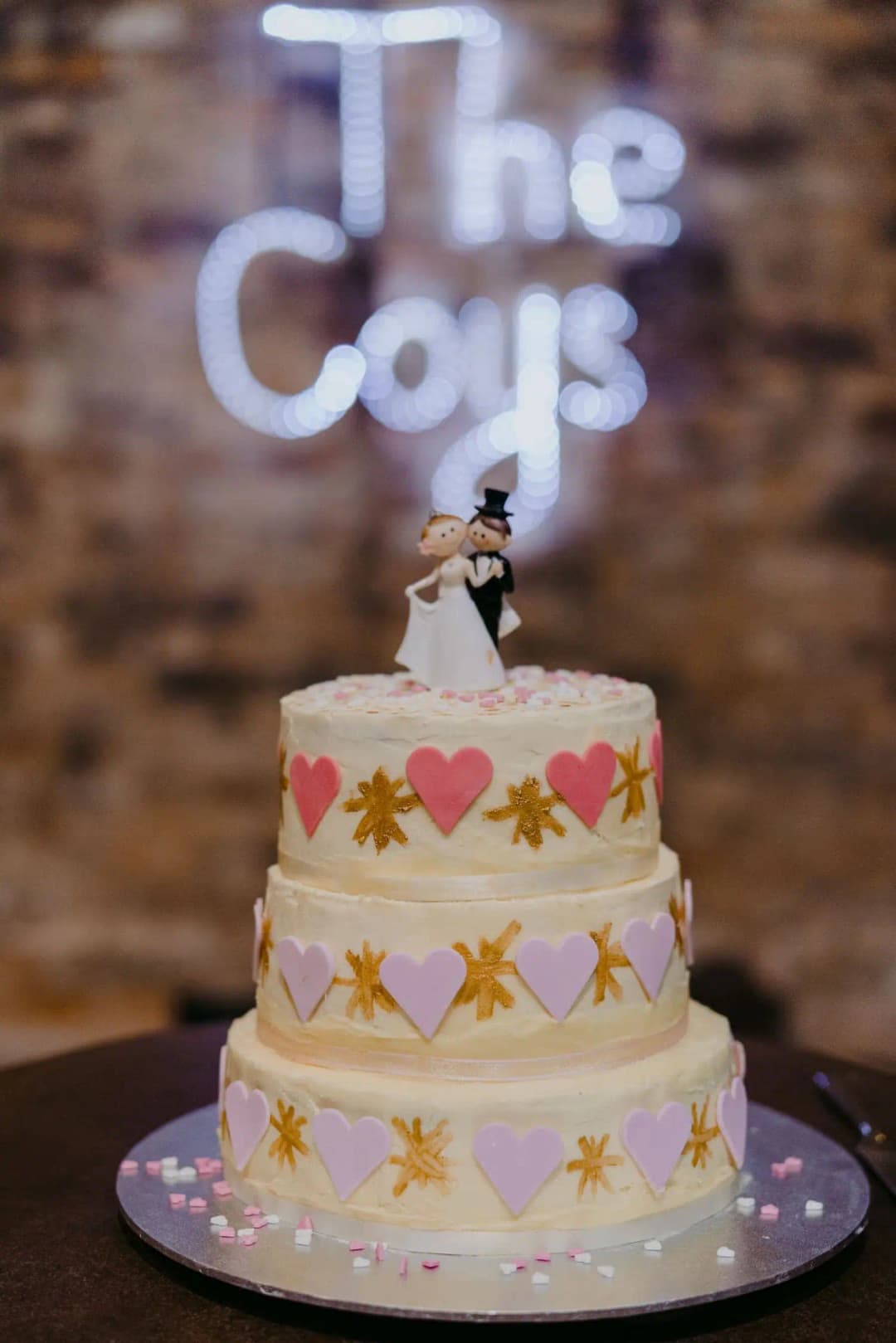 Three-tier wedding cake decorated with colourful hearts and leaves beneath an illuminated wall sign.