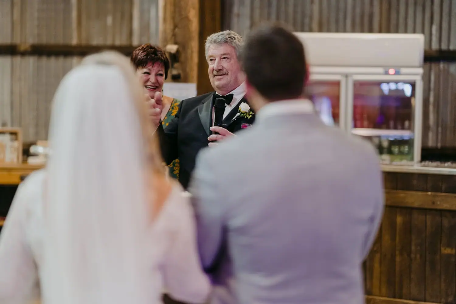 Man giving a microphone speech in front of the seated bride and groom.