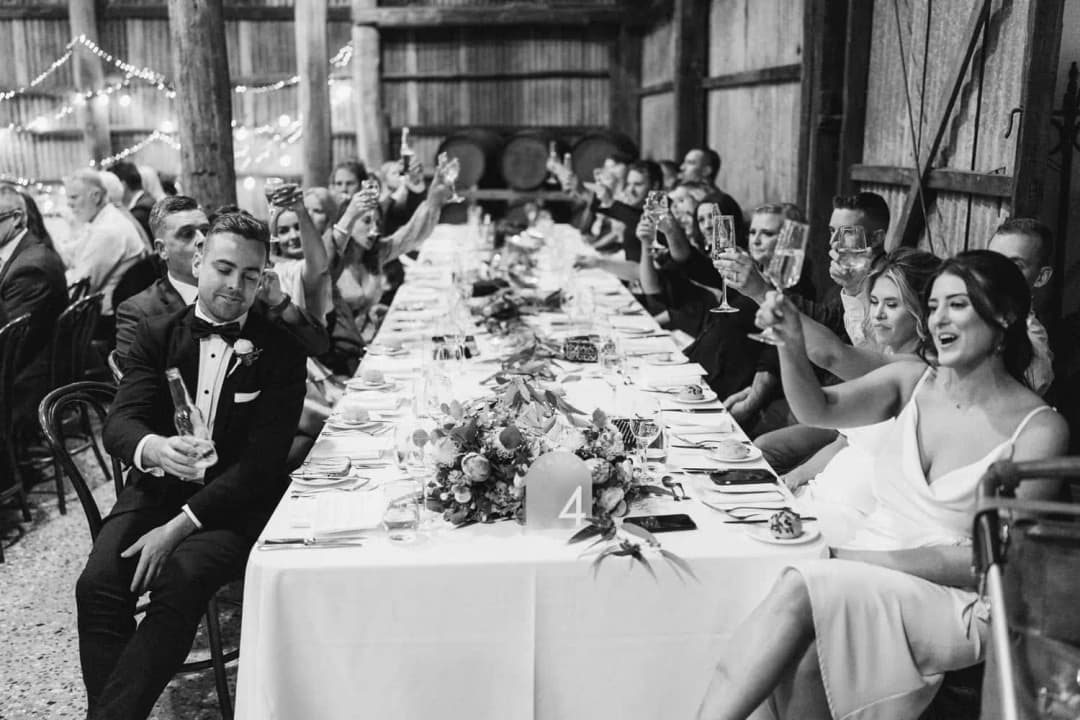 Black-and-white photograph of guests raising glasses along a long reception table.