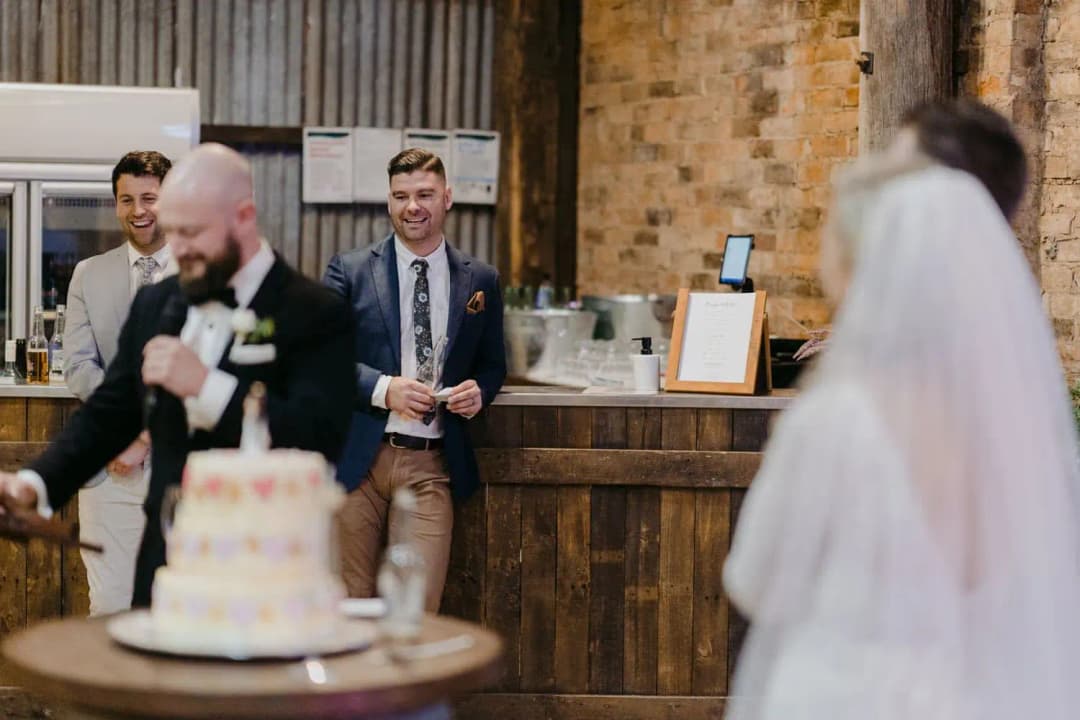 Man speaking into a microphone beside the wedding cake as the bride watches.