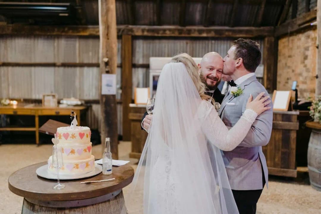 Bride and groom embracing a smiling man beside their wedding cake.