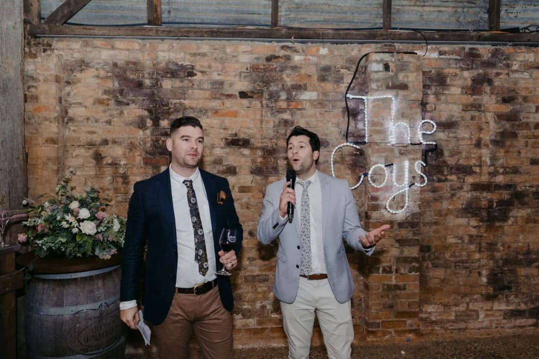 Two men speaking into microphones beneath an illuminated wall sign.