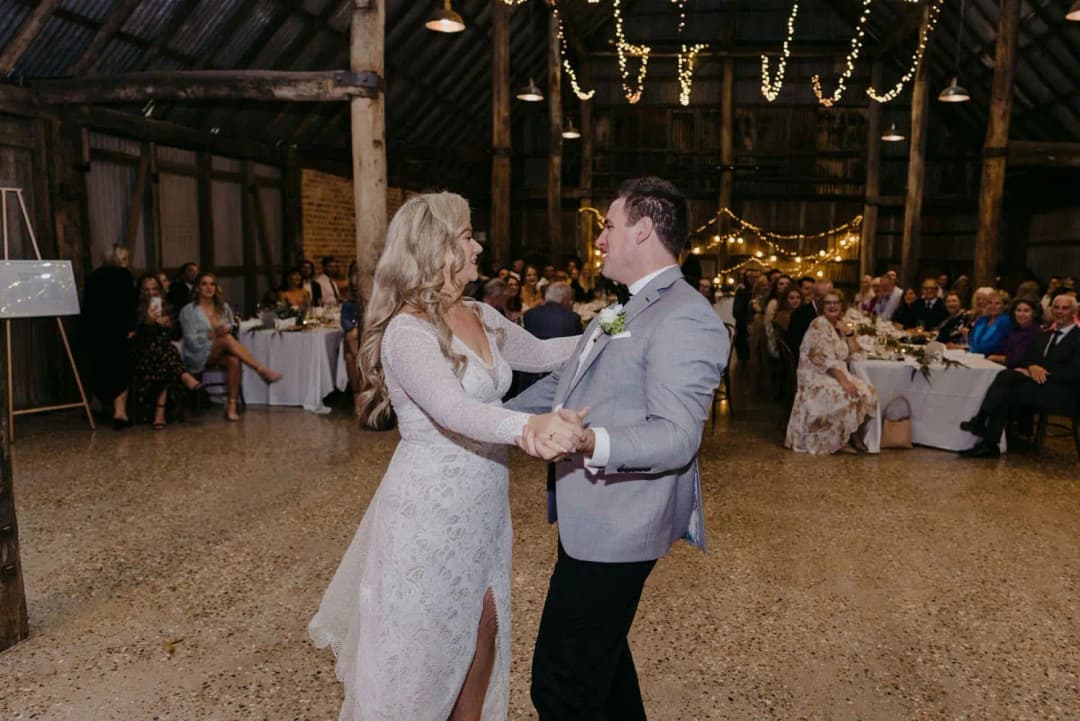 Bride and groom smiling while dancing beneath timber beams and string lights.