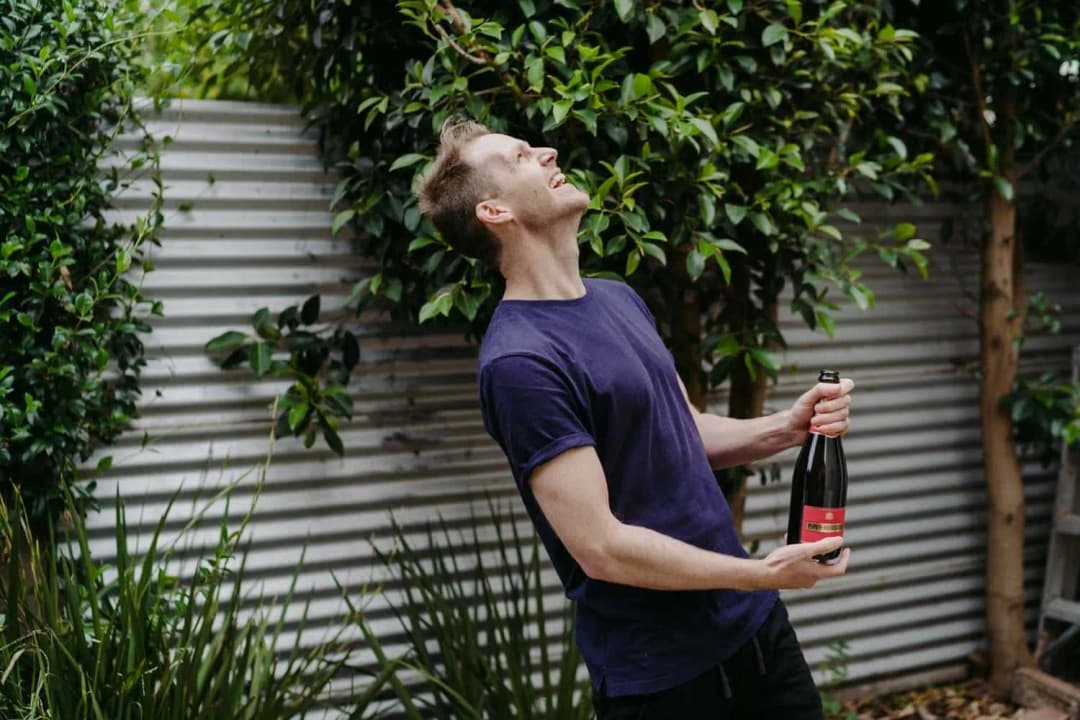Man looking upward while holding a bottle in a leafy backyard