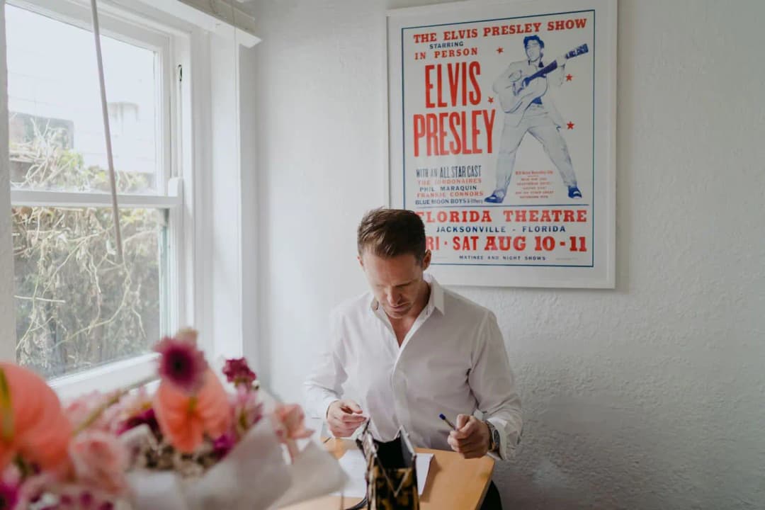 Groom holding a pen beside a window beneath an Elvis Presley poster