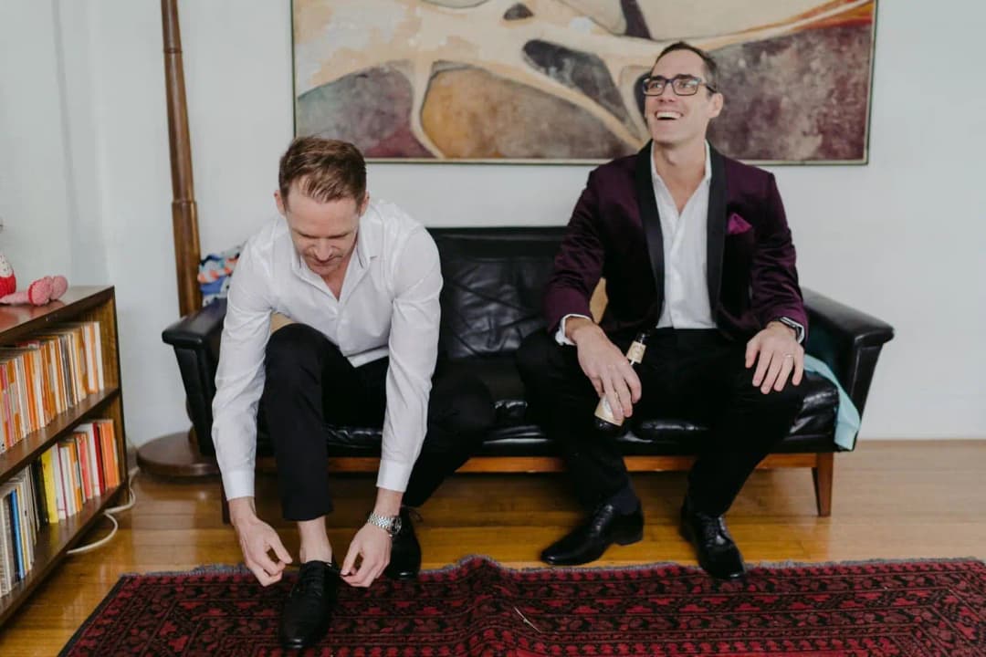 Two men laughing on a sofa while one fastens his shoes