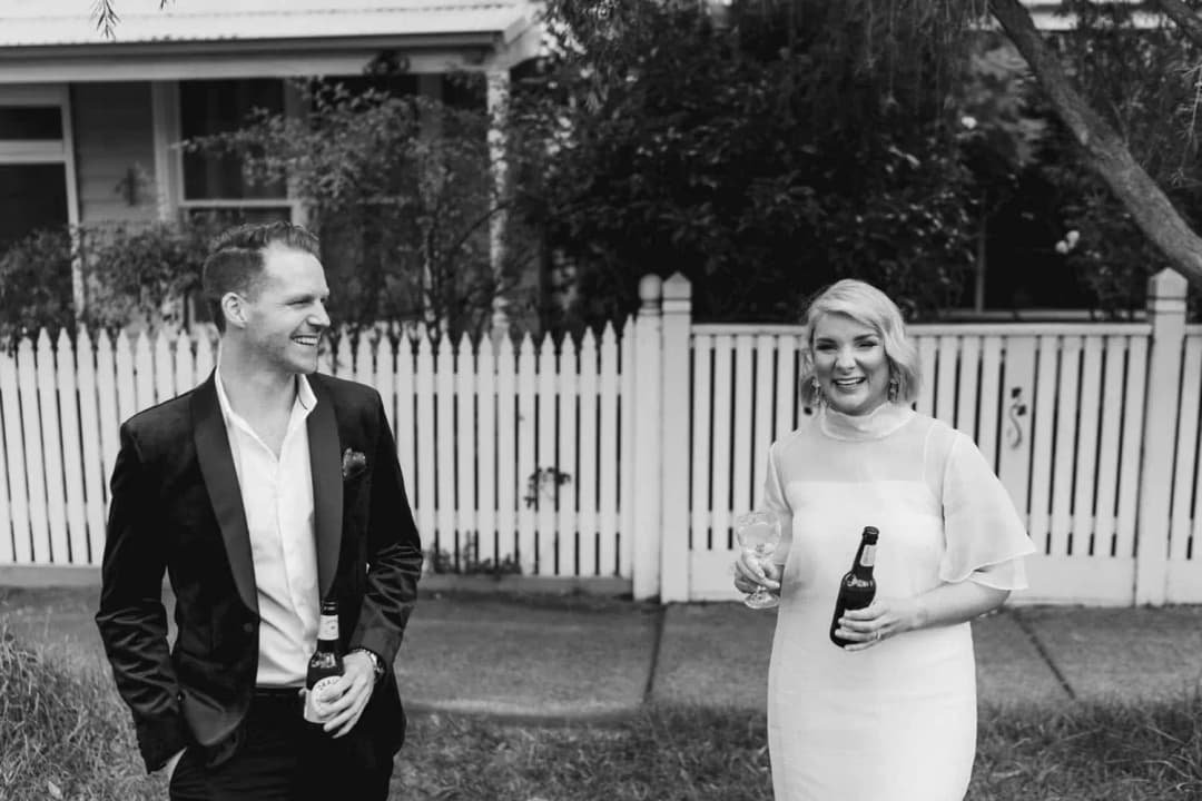 Black and white photograph of the couple smiling with bottles beside a picket fence