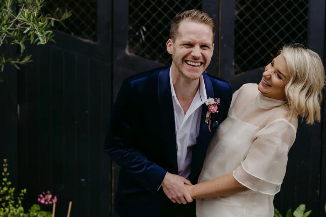 Bride and groom laughing together beside a dark doorway and leafy plants
