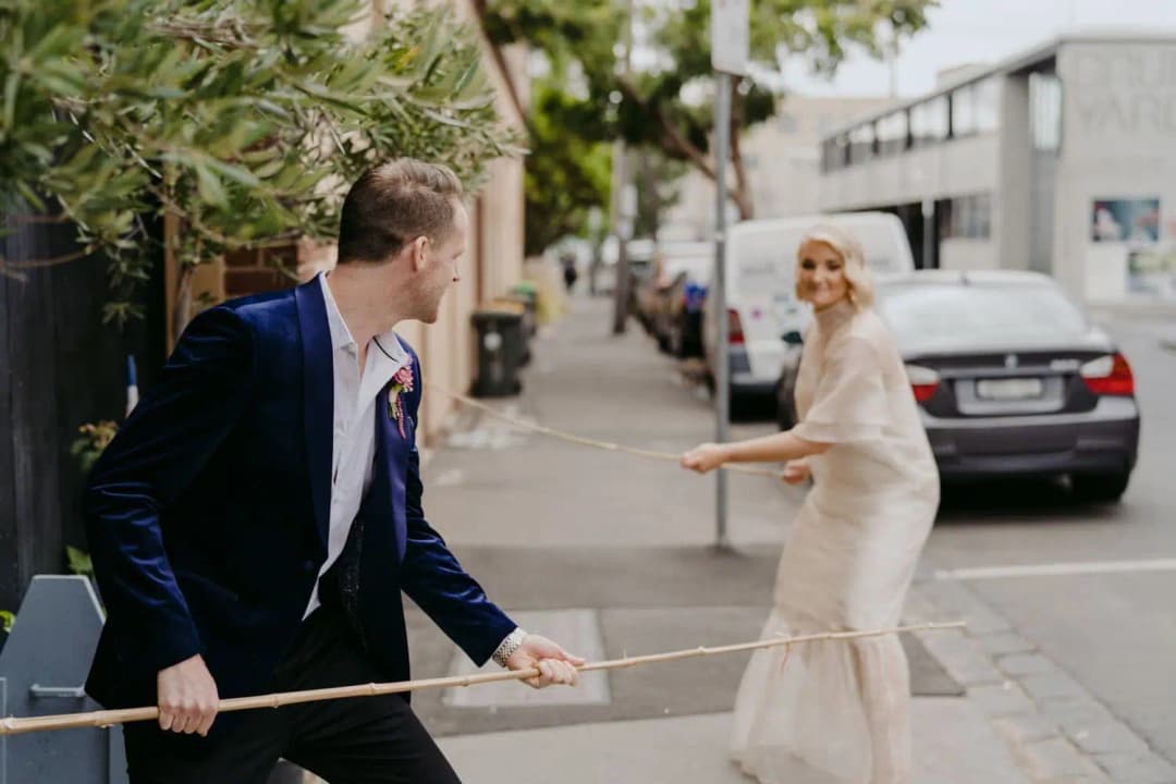 Bride and groom playfully facing each other with long bamboo poles on a footpath