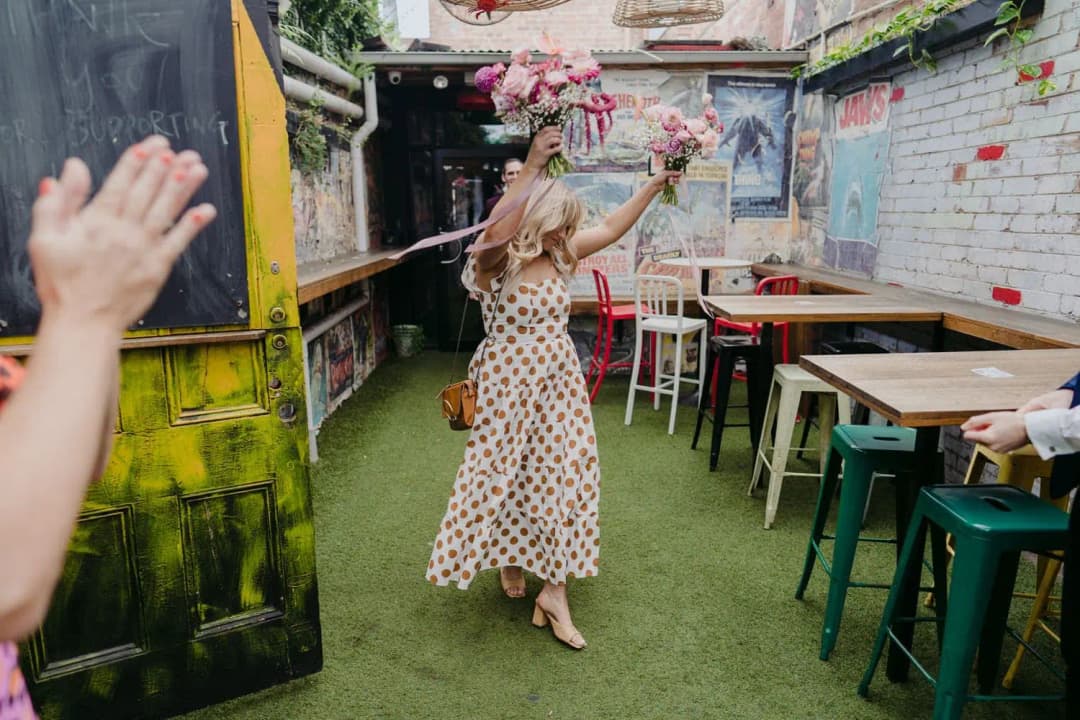 Bridesmaid raising her bouquet while entering a colourful brick courtyard