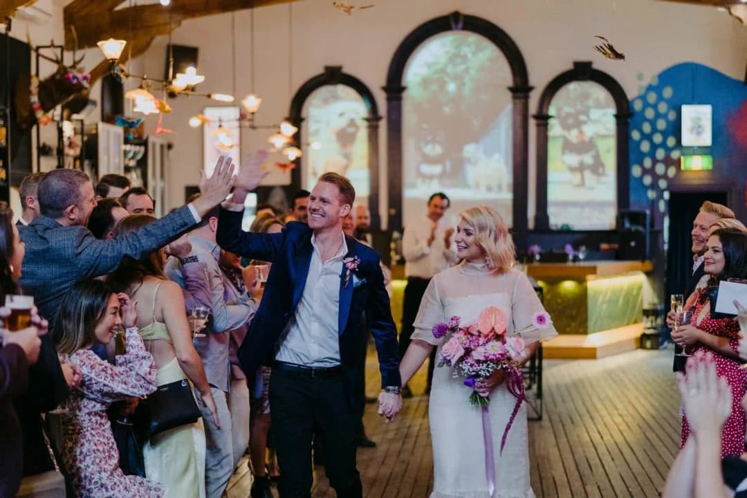 Groom greeting a guest with a raised hand while walking beside the bride