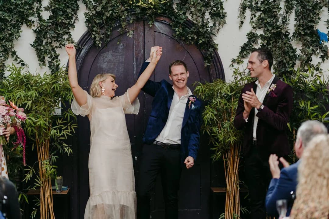 Bride raising both arms beside the groom as guests applaud