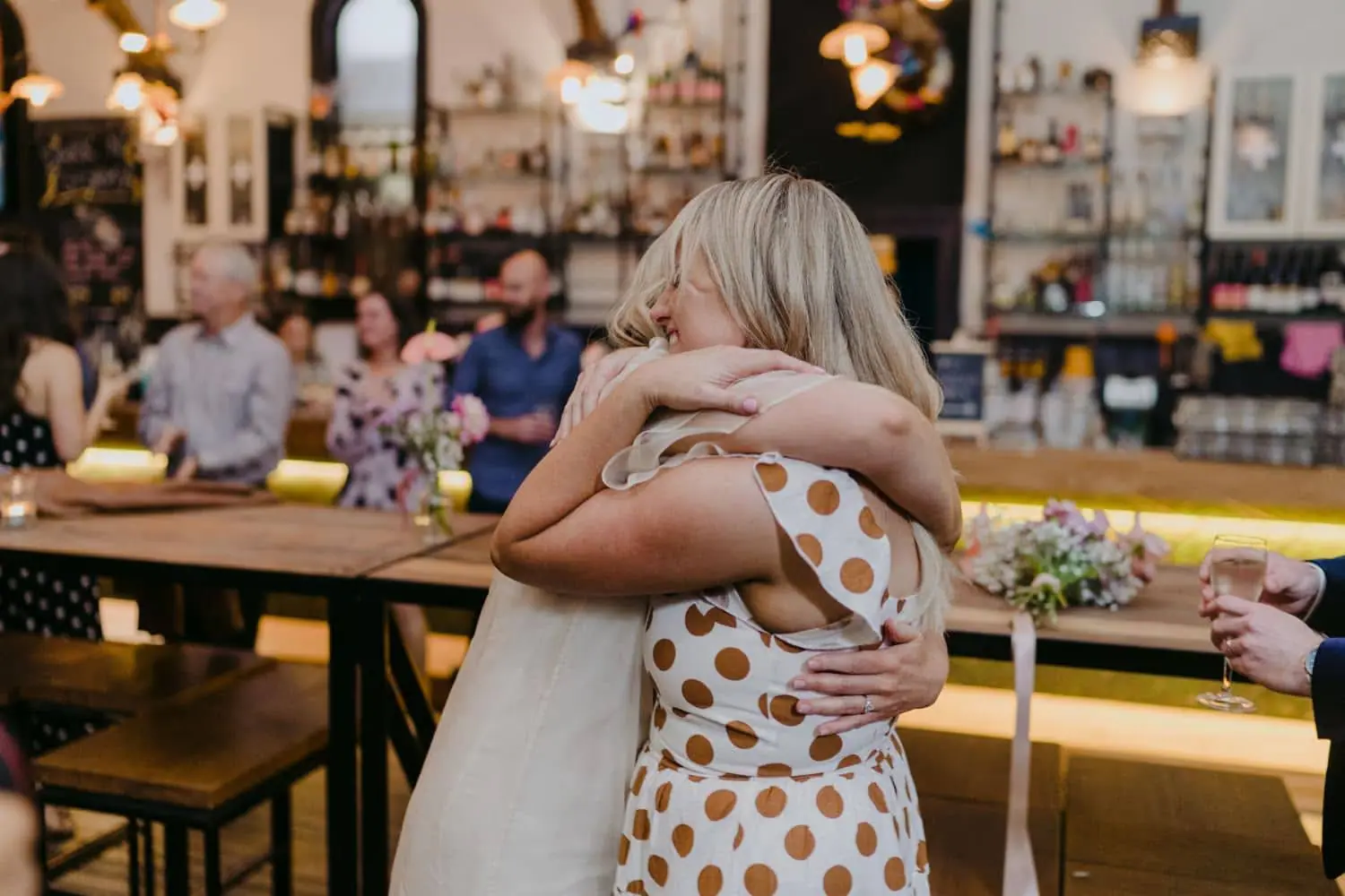 Bride hugging a woman in a white and gold spotted dress beside the bar