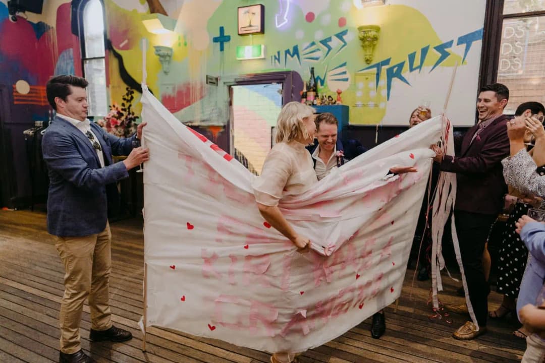 Bride leaning forward into a paper banner held up by wedding guests