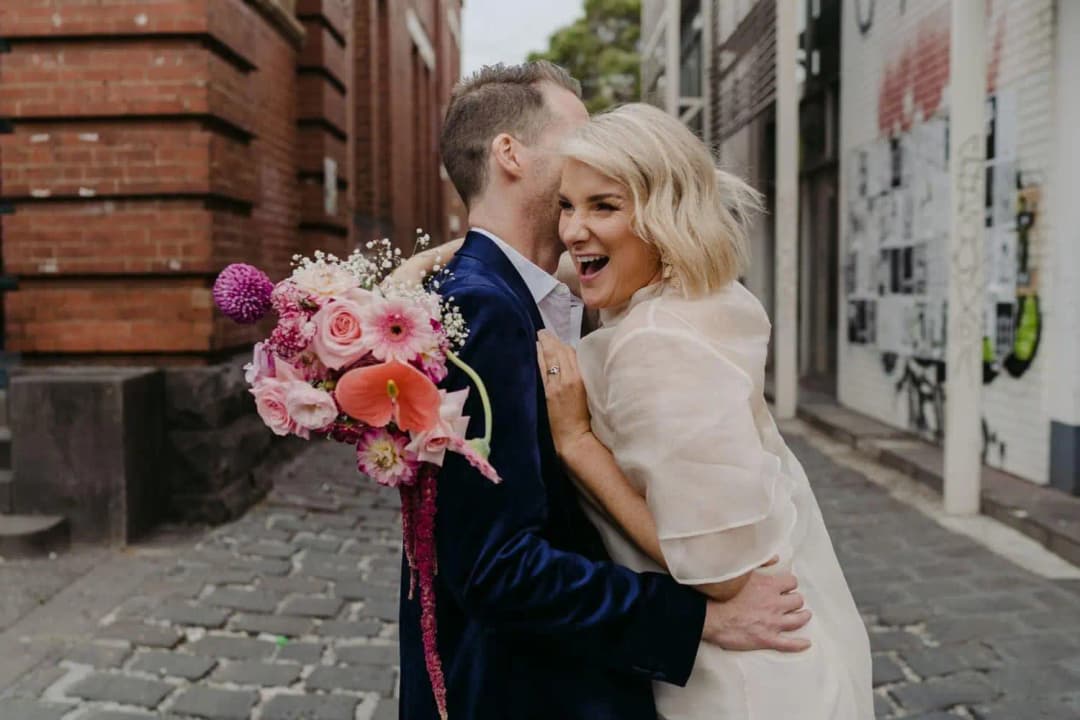 Bride laughing as the groom leans toward her beside a brick wall
