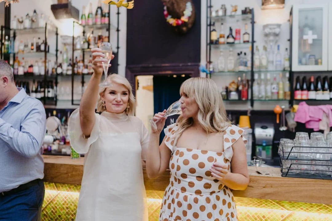 Bride raising her glass beside a smiling bridesmaid at the reception bar