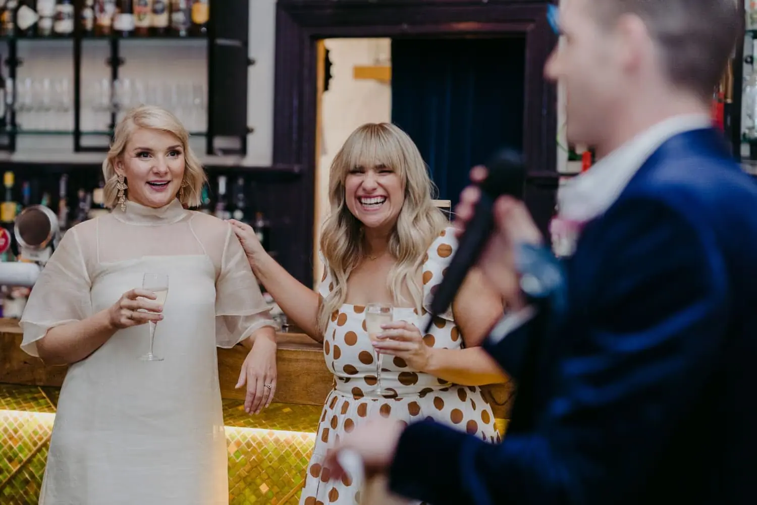 Bride and bridesmaid smiling as the groom speaks into a microphone