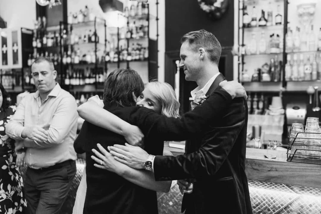 Black and white photograph of the couple embracing a guest beside the bar