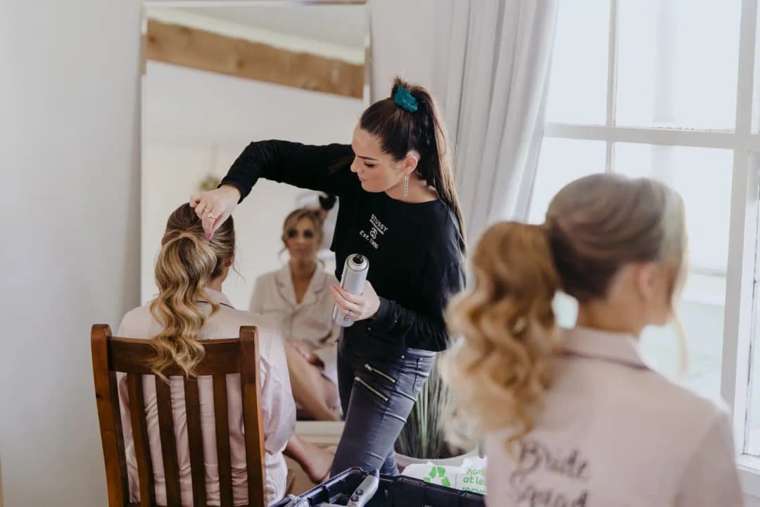 Hair stylist working on the bride’s hair beside a bedroom window