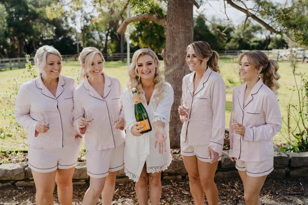 Bride holding a bottle as bridesmaids in pale pyjamas laugh outdoors