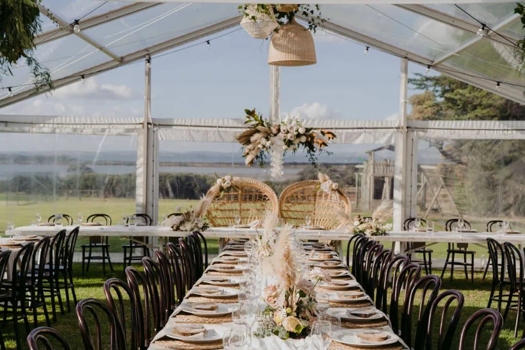 Long dining table beneath a clear marquee roof with two wicker chairs at its end